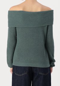 Maglione a spalla scoperta in verde tenue con texture a costine. Presenta maniche lunghe e una vestibilità ampia, abbinato a pantaloni in denim scuro.