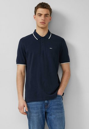 Młody mężczyzna oparty o ścianę, ubrany w granatową koszulkę polo z białym obszyciem i niebieskie jeansy, jedna ręka w kieszeni, neutralny wyraz twarzy.