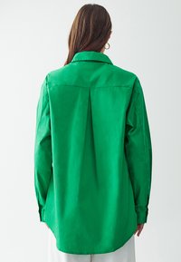 Vihreä pitkähihaista paitaa, jossa on kauluspää, takana pleat-yksityiskohta, valmistettu sileästä ja kevyestä kankaasta.