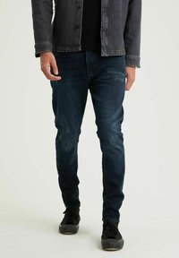 Mörkblå denimjeans med slim fit, med slitna detaljer och subtila blekningar. Bärs med en svart stickad tröja och en grå jacka.