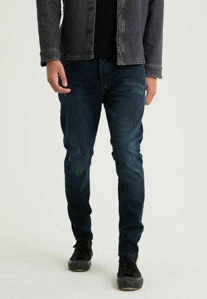 Dark blue denim jeans with a slim fit, featuring distressed accents and subtle fading. Worn with a black knit sweater and gray jacket.