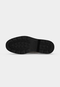 Semelle de chaussure noire avec des motifs de bande de roulement circulaires et rectangulaires, montrant une empreinte du nom de la marque dans la section centrale sur fond blanc.