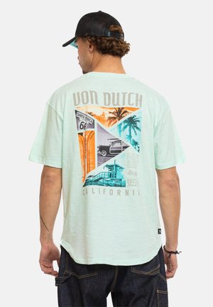 Homme portant un t-shirt vert clair Von Dutch avec des graphismes vintage de la Route 66 en Californie et un palmier dans le dos, associé à un jean foncé et une casquette noire.