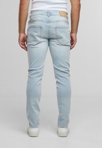 Vyras, stovintis nugara, vilkintis šviesiai mėlynus siaurus džinsus, baltus sportbačius ir baltą marškinį, prieš paprastą pilką foną.