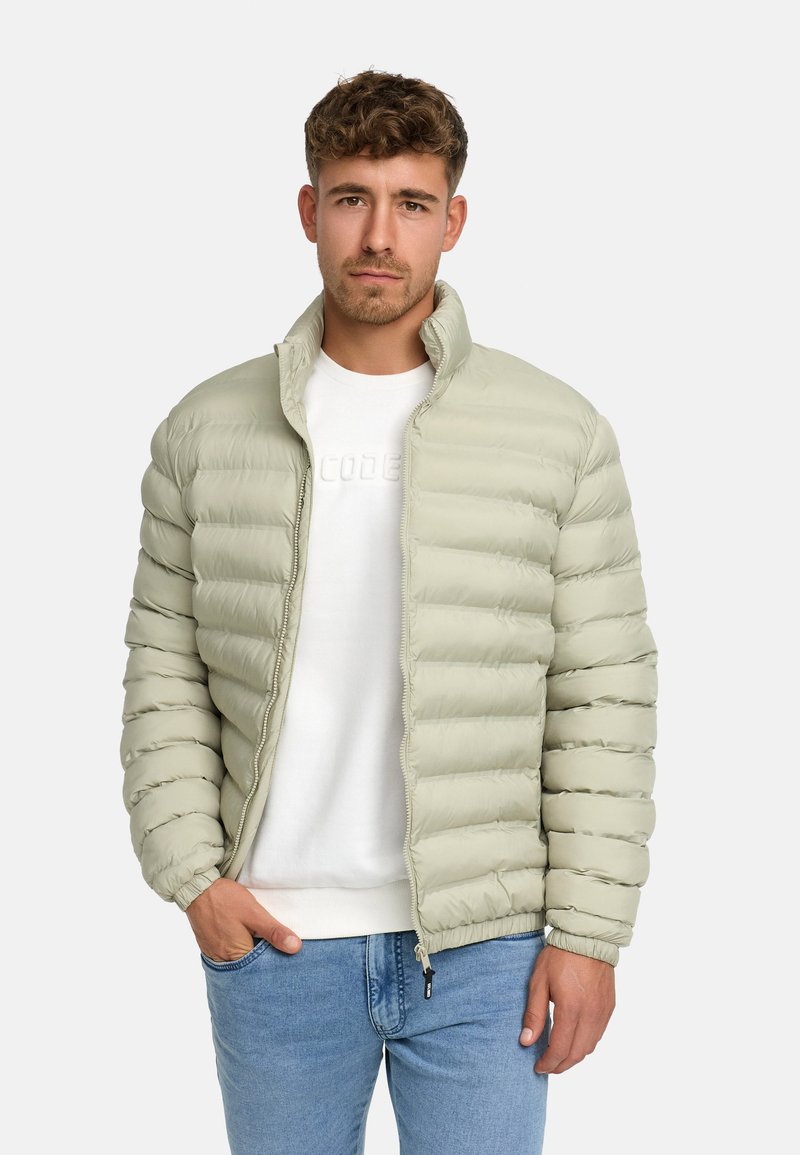 Lys grøn puff jakke med quiltede sektioner, fuld lynlås, høj krave og elastiske manchetter, båret over en hvid sweatshirt og blå jeans.