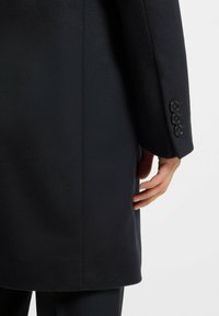 Cappotto di lana nera con una texture liscia, vestibilità sartoriale e dettagli a tre bottoni sui polsini. Mano visibile appoggiata sul lato del cappotto.
