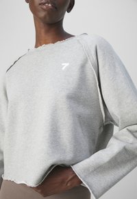 Personne à la peau foncée portant un sweat-shirt court à manches longues gris clair avec des bords bruts et un petit logo blanc "7" sur la poitrine.