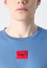 Modra bombažna majica s krožnim izrezom in rdečim pravokotnim obližem, na katerem je v črni barvi izvezen napis "HUGO". Gladka tekstura, sproščen dizajn.