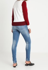 Suéter color bloque en rojo y blanco con mangas largas, combinado con jeans ajustados en azul claro y zapatos negros sin cordones. Textura suave, diseño ajustado.
