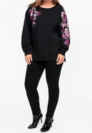 Femme portant un pantalon noir et un haut noir à manches longues avec des motifs floraux roses en sequins sur les épaules et les manches, assorti à des bottines noires à talons.