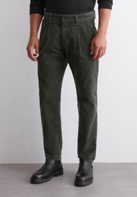 Tmavě zelené kordové kalhoty se střihy rovného nohavice, s předním zapínáním na knoflík a bočními kapsami, spárované s černými botami.