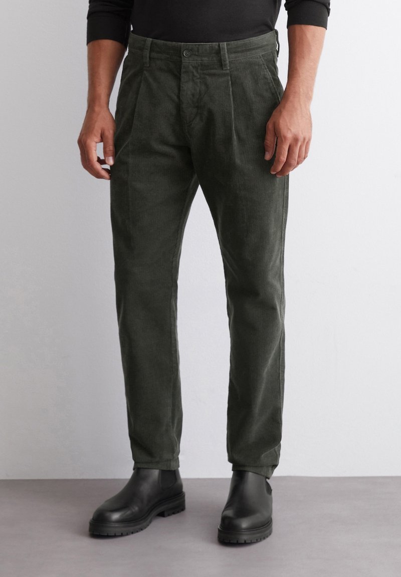 Tmavě zelené kordové kalhoty se střihy rovného nohavice, s předním zapínáním na knoflík a bočními kapsami, spárované s černými botami.
