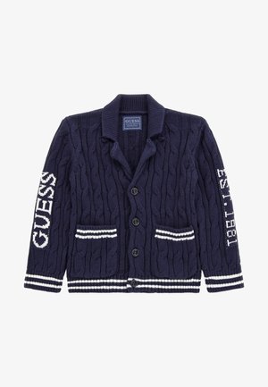 Cardigan bleu marine en maille torsadée avec boutons, poignets et poches à rayures blanches, inscription "GUESS" sur la manche gauche, et "EST. 1981" sur la manche droite.