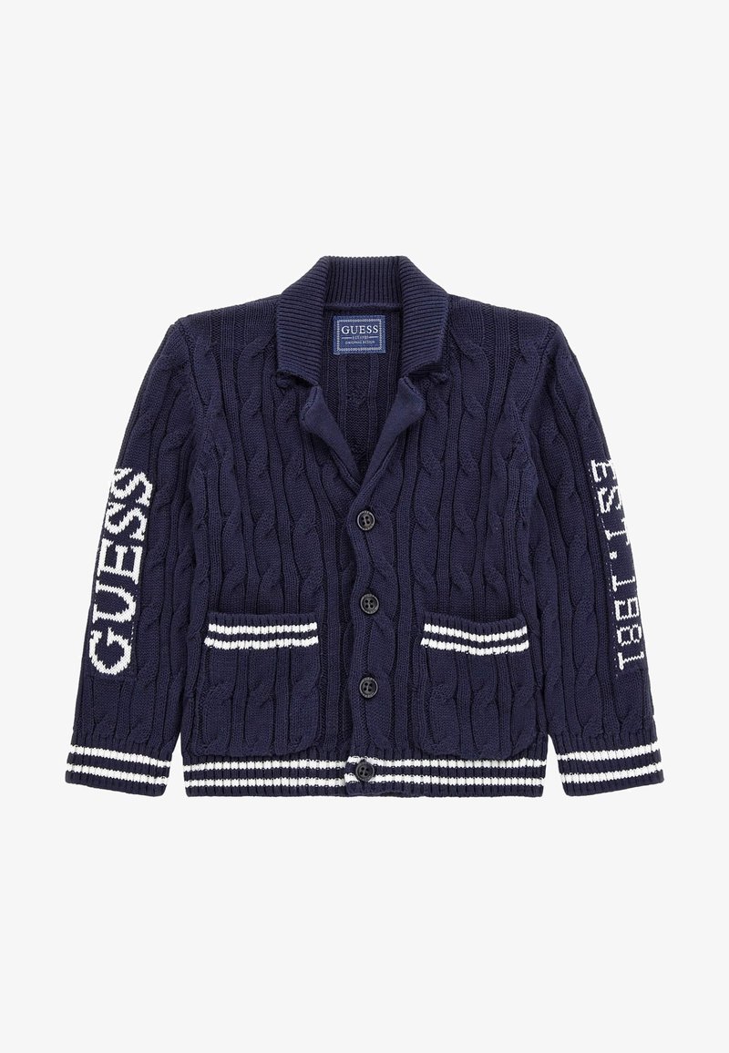 Cardigan bleu marine en maille torsadée avec boutons, poignets et poches à rayures blanches, inscription "GUESS" sur la manche gauche, et "EST. 1981" sur la manche droite.