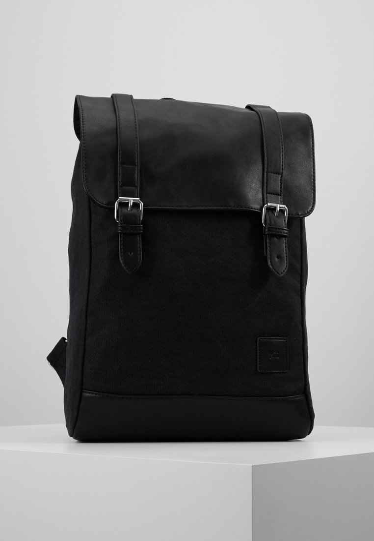Sac à dos noir avec un corps en tissu texturé et un rabat en cuir lisse. Comprend deux boucles en métal, une petite étiquette logo et une poche latérale.
