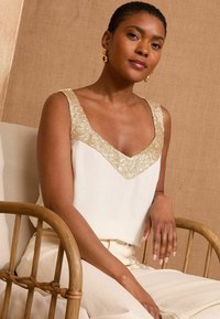 White sleeveless top with a gold sequin V-shaped neckline, paired with light-colored pants, worn while seated on a wooden chair.