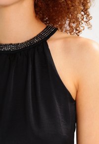 Svart ärmlös topp med en rynkad halsringning prydd med mörka, glänsande detaljer. Slät tyg med en mjuk textur.