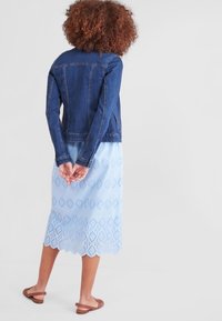 Jeansjack, blauwe mouwloze jurk met oogjes en bruine sandalen. De jurk heeft een gekartelde zoom en diamantpatronen.