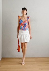 Haut sans manches coloré et à motifs avec des bretelles volantées, associé à une jupe blanche et un sac à main rouge. Modèle portant des sandales à lanières.