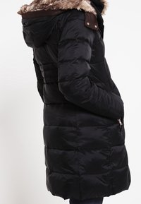 Svart puffjacka med en luva av fuskpäls, dragkedjefickor och en kviltad textur. Har en mörkbrun accent vid kragen.