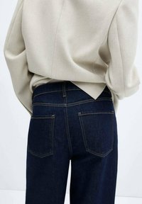 Personne portant un jean taille haute bleu marine avec des poches arrière et un haut beige à manches longues rentré à la taille.