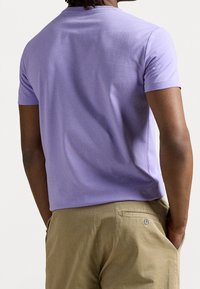 Man som har på sig en åtsittande lavendelfärgad kortärmad skjorta och beigefärgade byxor med händerna i fickorna, sedd bakifrån.