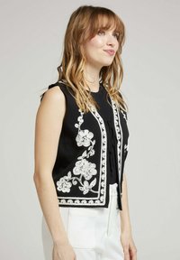 Gilet sans manches noir avec des broderies florales blanches le long des bords ; présente un tissu texturé et un design ouvert à l'avant.
