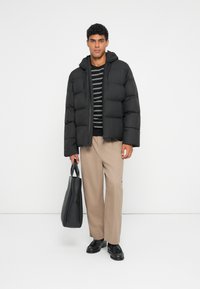 Giacca a vento nera con cappuccio, chiusura con zip e design trapuntato. Indossata sopra una maglietta a righe, abbinata a pantaloni color beige e una borsa nera.