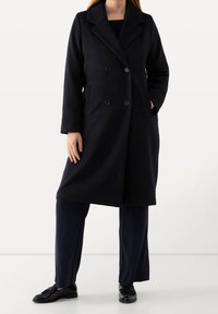 Cappotto nero doppiopetto in misto lana, con ampi revers, due tasche frontali e taglio dritto. Indossato con pantaloni blu navy e scarpe.