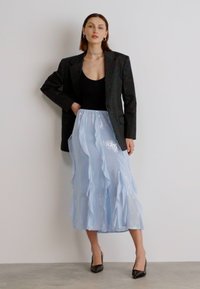 Blazer noir, body noir, jupe midi plissée bleu clair avec volants, texture brillante, escarpins noirs, sur fond neutre.