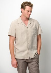 Camicia a maniche corte beige chiara in lino con colletto classico, taschino singolo sul petto e vestibilità rilassata, abbinata a pantaloni marrone chiaro.