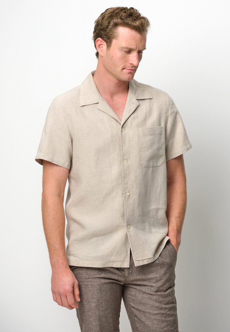 Camicia a maniche corte beige chiara in lino con colletto classico, taschino singolo sul petto e vestibilità rilassata, abbinata a pantaloni marrone chiaro.