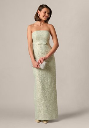 Vrouw in een strapless, lichtgroene jurk met structuur, die een zilveren clutch vasthoudt, glimlacht en staat tegen een effen achtergrond.