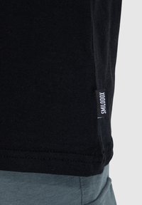 T-shirt nera in cotone con maniche corte e un'etichetta laterale che mostra "SMILODOX". Tessuto morbido e design semplice. Tessuto grigio visibile sotto.