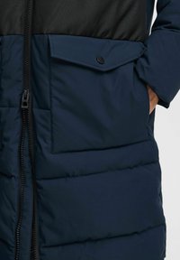 La chaqueta puffer de color azul marino oscuro cuenta con un cierre de cremallera, un gran bolsillo en el pecho y un tejido negro texturizado en la mitad superior. Material suave e insulated.