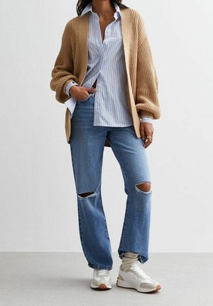 Osoba ubrana w luźny beżowy sweter na drutach, niebieską koszulę w paski zapinaną na guziki, podarte niebieskie dżinsy, beżowe skarpetki i białe tenisówki, stojąca z jedną ręką w kieszeni.