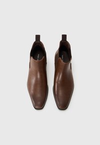 Botas de piel marrón hasta el tobillo con un acabado suave, inserciones laterales elásticas y tiradores para facilitar su puesta. Presentan una punta afilada y suela plana.