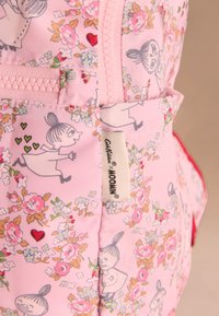 Rózsaszín szövet hátizsák virágmintával és rajzfilmfigurákkal, cipzáras záródással és márkázott címkével, semleges háttér előtt elhelyezve.