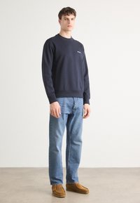 Sudadera azul marino con cuello redondo, puños y dobladillo acanalados. Combinada con jeans rectos de color azul claro y zapatos estilo mocasín marrones.