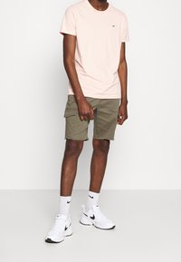 Muž oblečený v světle růžovém tričku, olivově zelených cargo šortkách, bílých ponožkách Nike a bílých teniskách Nike stojí proti jednobarevnému pozadí.