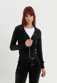 Zwarte fluwelen hoodie met rits en een trekkoord, met een aansluitende pasvorm, geribde mouwen en een kangoeroezak. Draagt over een witte top.