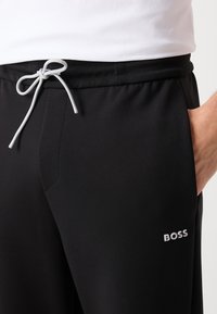 Svarta joggingbyxor gjorda av mjukt material med elastisk midja, justerbar dragsko, sidfickor och en vit "BOSS"-logotyp.