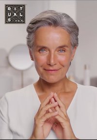 Une femme aux cheveux gris courts et aux yeux bleus porte un haut blanc, les mains jointes devant elle. Un éclairage doux et naturel met en valeur ses traits.
