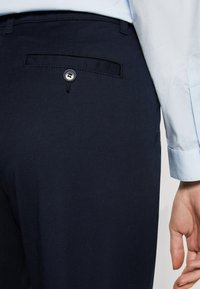 Námornícke nohavice s zadným vreckom so zapínaním na gombík. Tkanina vyzerá hladko, so strihom na mieru, ktorý je zvýraznený svetlo modrým rukávom košele.