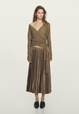 Un pull en olive, style portefeuille avec des manches longues, associé à une jupe longue plissée en bronze métallique. Des ballerines noires à bout pointu.