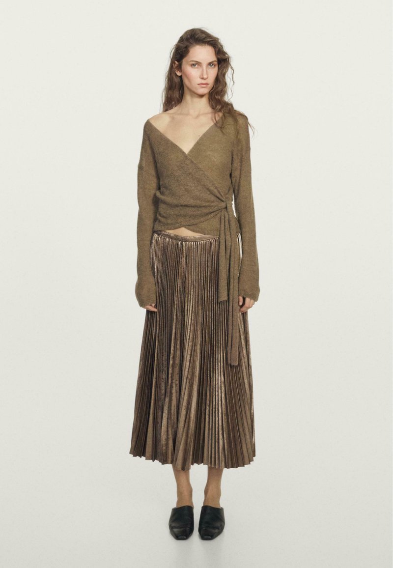Un pull en olive, style portefeuille avec des manches longues, associé à une jupe longue plissée en bronze métallique. Des ballerines noires à bout pointu.