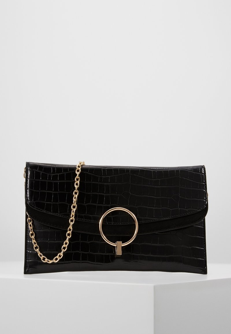 Pochette noire à texture crocodile avec fermoir en anneau circulaire doré et bandoulière chaîne dorée attachée sur une surface blanche.
