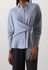 Camicia a righe blu e bianche con design avvolgente. Presenta maniche lunghe e un orlo curvo. Il tessuto testurizzato aggiunge una sottile profondità.