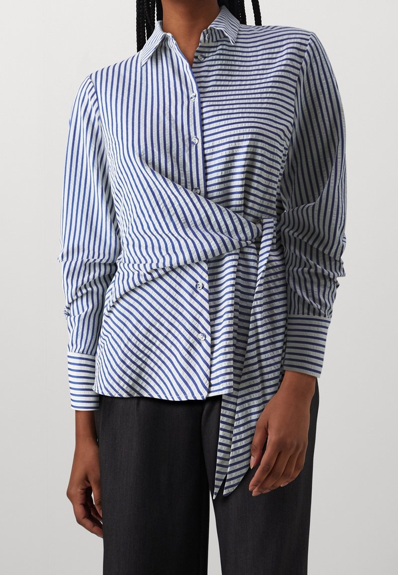 Camicia a righe blu e bianche con design avvolgente. Presenta maniche lunghe e un orlo curvo. Il tessuto testurizzato aggiunge una sottile profondità.