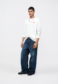 Camisola branca de manga longa com o logotipo vermelho "DIESEL", combinada com uns jeans largos de cor azul escura e sapatos pretos; modelo apresenta-se sobre um fundo simples.